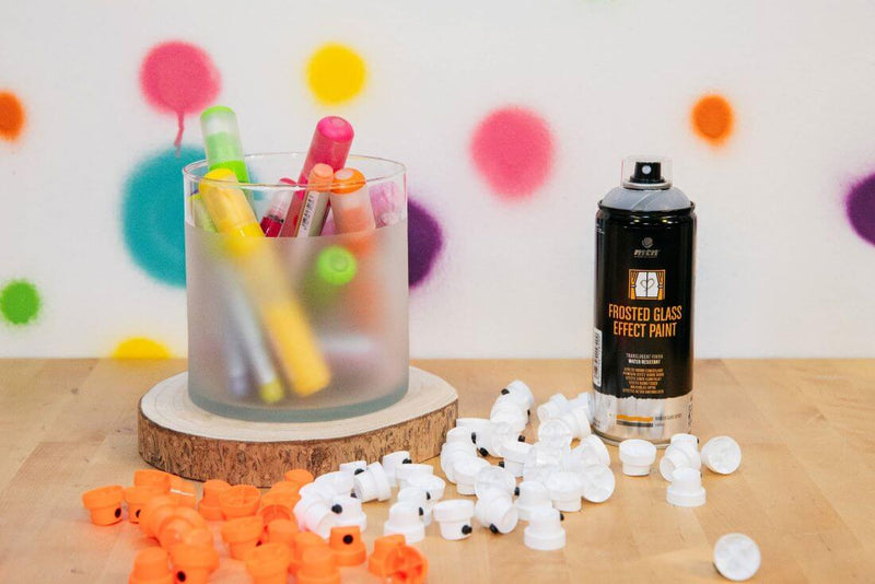 Lata de Spray MTN PRO Frosted Glass Paint junto a um jarro pintado pelo mesmo, com marcadores coloridos no seu interior. Junto a ambos os objetos encontram-se vários bicos Montana, brancos e laranjas.