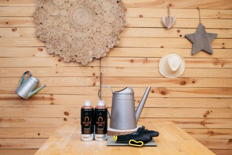 Duas latas de Spray Montana Efeito Mármore pousadas sobre uma mesa de madeira, junto a uma caneca de metal, antes de ser pintada.